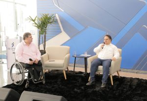 Foto de palestrantes durante evento. São dois homens. Um dos homens senta em uma poltrona e fala em um microfone de mão. O outro está sentado na cadeira de rodas e segura um microfone no colo.