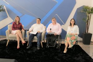 Foto de palestrantes do evento. Na foto há 2 mulheres e 2 homens sentados em assentos individuais. Um dos homens segura um microfone de mão.