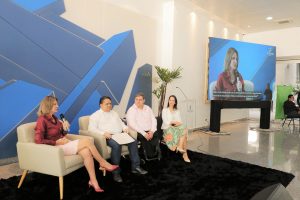 Foto de palestrantes do evento. Na foto há 2 mulheres e 2 homens sentados em assentos individuais. Uma das mulheres fala em um microfone de mão. A direita do grupo há um telão, transmitindo os palestrantes em alta resolução.