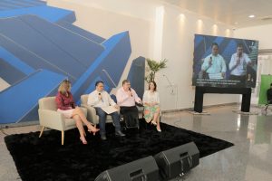 Foto de palestrantes do evento. Na foto há 2 mulheres e 2 homens sentados em assentos individuais. Ambos os homens falam em microfones de mão. A direita do grupo há um telão, transmitindo os palestrantes em alta resolução.