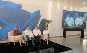Foto de palestrantes do evento. Na foto há 2 mulheres e 2 homens sentados em assentos individuais. Um dos homens fala em um microfone de mão. A direita do grupo há um telão, transmitindo os palestrantes em alta resolução.