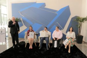 Foto dos participantes e palestrantes do evento. Na foto há 2 mulheres e 2 homens sentados em assentos individuais, a esquerda deles há um homem de pé falando no microfone. Estão todos posicionados lado a lado.