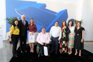 Foto dos participantes e palestrantes do evento reunidos para foto. Na foto há 5 mulheres e 2 homens de pé, e um cadeirante. Todos alinhados lado a lado posando para a foto.