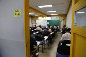 Foto colorida mostrando, de costas, os candidatos do concurso sentados em carteiras fazendo as provas 