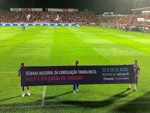 Foto colorida mostra jogadores no gramado do campo do Vila Nova segurando faixa com texto sobre a Semana Nacional da Conciliação Trabalhista, no intervalo do jogo do Campeonato Brasileiro de Futebol