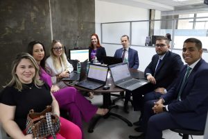 Foto de representantes da Equatorial e das partes autoras das ações em audiência com a conciliadora no Cejusc de Goiânia. Na foto, 4 mulheres e 3 homens estão sentados ao redor de uma mesa redonda, posando e sorrindo para a foto. Sobre a mesa há notebooks.