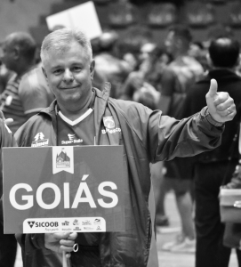 Foto em preto e branco do Servidor Fernando Prata nas Olimpíadas da Justiça do Trabalho em 2018. Trata-se de um homem branco, de cabelo liso, curto e grisalho. Ele veste uma jaqueta de frio. Está segurando uma placa que tem escrita em si a palavra "goiás" e posa para a foto fazendo um sinal de "joia" com a outra mão.