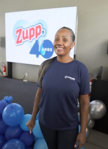 Foto da operadora de máquinas, Carla Silva. Trata-se de uma mulher negra de cabelo liso, grisalho e amarrado em um rabo de cavalo. A mulher está vestindo uma camiseta azul escura e calça preta. Ela está sorrindo enquanto posa para a foto. 