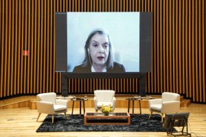 Foto colorida da ministra Cármen Lúcia aparecendo em um telão no fundo do palco do auditório Goyazes. Trata-se de uma mulher branca com cabelo liso e grisalho na altura do ombro. A mulher veste blazer preto e utiliza um fone de ouvido com fio branco. À frente, no palco, estão dispostas três poltronas, duas mesas laterais e uma mesa ao centro.