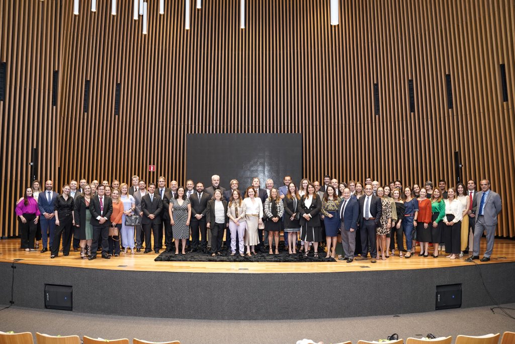 Foto de um grande grupo de pessoas posando para uma foto no palco do auditório Goyazes do TRT-GO. Elas estão de pé, posicionadas lado a lado em duas fileiras que se estendem pelo palco. 