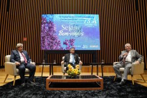 Foto de três desembargadores durante ocorrência do evento. Eles estão sentados ao redor de uma mesa central, em três poltronas brancas dispostas no palco. Estão vestidos formalmente, com terno e se direcionam para a plateia. Atrás dos homens há um grande telão projetando o texto "Sejam bem-vindos". 