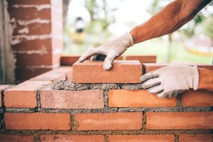 Foto de um trabalhador assentando tijolos em uma construção. Na imagem, há o recorte de duas mãos vestidas com luvas de borracha e assentando tijolos