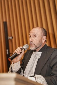 Foto de Alpiniano Lopes, chefe do MPT-GO, durante cerimônia. Trata-se de um homem branco e calvo, está vestido de toga preta. Ele está segurando um microfone de mão sem fio e discursando atrás de um púlpito de madeira. 