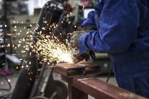 Foto colorida mostra homem soldando barra de ferro. Na imagem, o tronco de um homem vestido de macacão azul e luvas pretas utiliza a ferramenta de solda numa coluna metal apoiada em um toco de madeira.