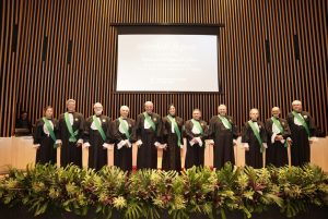 Foto dos desembargadores presentes na cerimônia. Estão todos vestidos de toga preta sobreposta por uma faixa verde com comenda. Tratam-se de duas mulheres e nove homens, todos posicionados lado a lado em cima do palco do auditório dos Goyazes. Eles posam e sorriem para a foto. No fundo há um grande telão de projeção branco.