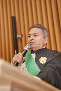 Foto de Geraldo Nascimento, presidente do TRT-18, durante cerimônia. Trata-se de um homem branco, de cabelo curto, liso e grisalho, está vestido com uma toga preta sobreposta por uma faixa verde. Ele está segurando um microfone de mão sem fio e discursando atrás de um púlpito de madeira. 