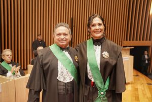 Foto do desembargador e presidente do TRT-18, Geraldo Nascimento, e da desembargadora Wanda Lúcia durante cerimônia. O desembargador é um homem branco, de cabelo curto, liso e grisalho. A desembargadora é uma mulher branca, de cabelo liso e preto, amarrado num coque baixo com a franja solta. Ambos vestem uma toga preta sobreposta com uma faixa verde. Eles estão se abraçando de lado e sorrindo para a foto.