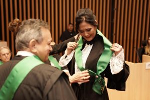 Foto da desembargadora Wanda Lúcia recebendo faixa e comenda do presidente do TRT-18, desembargador Geraldo Nascimento. A mulher branca, de cabelo liso, preto, preso em um coque baixo com a franja solta, está vestida de toga preta enquanto veste a faixa de cor verde entregue pelo presidente. O presidente, também vestido com a toga, aparece de costas para a foto e de frente para a desembargadora.