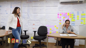 Foto colorida mostra dinâmica durante oficina. Na imagem, uma mulher está sentada atrás de uma mesa de madeira, e outra está de pé, a sua esquerda. No fundo, há uma parede cheia de post its pregados em uma parede branca.