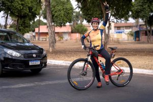 Foto mostra mulher sorrindo em pé atrás de uma bicicleta com o braço esquerdo levantado e a mão formando um "v" com os dedos indicador e médio