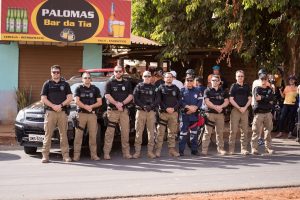 Foto colorida mostra oito homens usando calça ocre e camisa preta com as mãos cruzadas em frente ao corpo. Há também um homem vestindo um uniforme azul de bombeiro