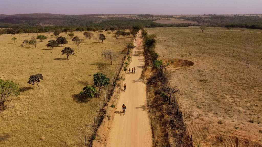 Imagem em orientação paisagem tirada por um drone mostra ciclistas pedalando em estrada de terra em via entre terrenos com grama baixa e algumas árvores à esquerda