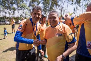 Foto mostra dois homens lado a lado em aperto de mãos. Ao fundo, árvores e céu aberto