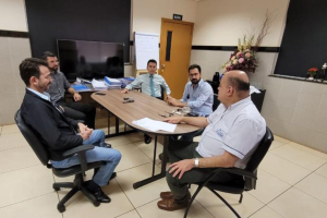 Foto colorida mostrando cinco homens sentados ao redor de uma mesa retangular.