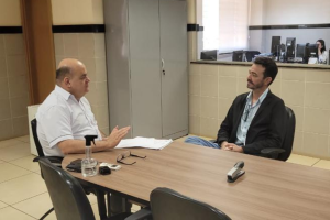 Em uma sala, dois homens sentados à mesa para uma reunião. O homem à esquerda usa camisa clara e o homem à direita usa um paletó azul marinho com uma camisa azul-claro.