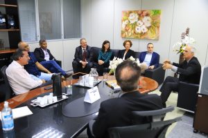 Foto colorida mostrando nove pessoas reunidas em uma sala. Tratam-se de duas mulheres e oito homens sentados em círculo.