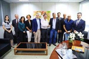 Foto colorida mostrando 11 pessoas vestidas com roupas formais. Tratam-se de três mulheres e oito homens sorrindo. Ao centro, dois homens apertam as mãos.