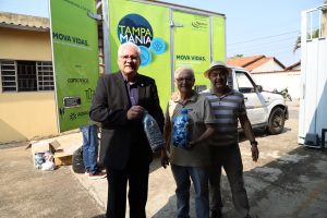 Em primeiro plano, três homens segurando garrafas plásticas com tampas de garrafas. Ao fundo, um caminhão baú com a cor verde escrito Tampamania