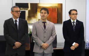 Três homens em pé com um quadro ao fundo. Uma figura abstrata nas cores marrom, amarelo e laranja