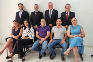 9 pessoas na foto. Em primeiro plano, tres mulheres e dois homens sentados com roupas coloridas. Atrás deles, 4 homens em pé, todos com terno e gravata, de tons escuros.