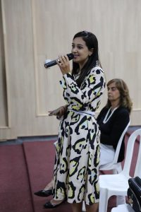 Foto da presidente da Comissão de Direito do Trabalho da Seccional de Anápolis, Evellyn Clemente, falando durante a reunião. Trata-se de uma mulher branca, de cabelo liso e preto longo. Ela usa um vestido branco com estampa de onça preta e amarela. A mulher falar em um microfone de mão. 