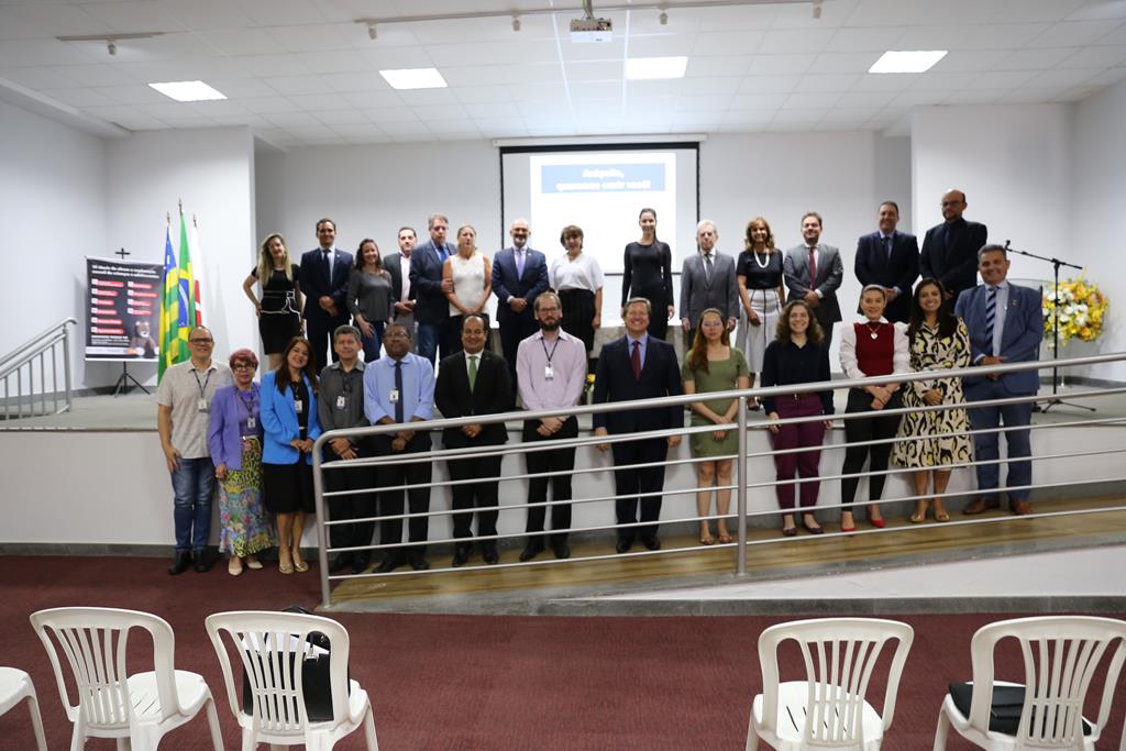 Foto de magistrados, juízes e advogados presentes na reunião. Na imagem, um grupo de pessoas constituído por homens e mulheres se posicionam lado a lado em duas filas, de pé, posando para a foto. Alguns estão em cima de um palco e outros em uma rampa conectada ao palco, na frente do mesmo.