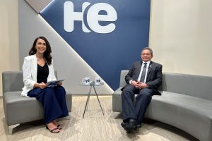 Foto de duas pessoas sentadas, uma mulher e um homem em dois sofás distintos, com uma pequena mesa ao centro. Em cima da mesa, duas canecas com a logo do HE. Ao fundo, um painel com a logo do HE