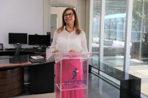 Foto colorida da juíza Eunice Castro discursando. Trata-se de uma mulher branca, de cabelo liso e loiro na altura do ombro. A juíza veste uma blusa de manga longa branca, saia longa rosa e óculos de armação preta. A mesma está falando atrás de um púlpito. 
