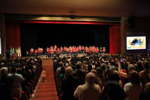 Foto mostra o auditório cheio e, no palco, integrantes da Banda Sinfônica