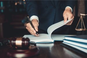 Foto das mãos de um homem usando uma caneta tinteiro para assinar um caderno. O caderno está em cima de uma mesa de madeira, posicionado ao lado de um martelo de juiz.