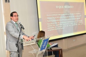 Foto mostra um homem de cabelos escuros, usando terno cinza e óculos de armação preta.  Ele está de pé, em frente a um púlpito, falando a um microfone e, ao seu lado, uma tela com um slide projetado. Há também uma moça sentada ao fundo.