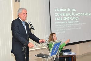 Foto mostra um homem grisalho de terno preto. Ele está de pé, em frente a um púlpito, falando a um microfone e, ao seu lado, uma tela com um slide projetado. Há também uma moça sentada ao fundo.