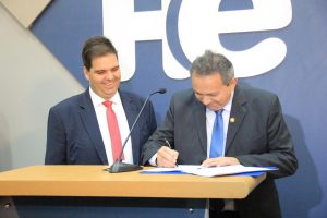 Foto colorida dos presidentes do TRT-18, desembargador Geraldo Nascimento, e da ABC, Reginaldo Júnior. Os dois homens estão atrás de um púlpito. O desembargador Geraldo Nascimento está à direita e tem pele clara, cabelos grisalhos, usa paletó preto, camisa social branca e gravata azul e está sorrindo enquanto assina um documento. Reginaldo Júnior está à esquerda e tem pele clara, cabelos escuros, está sorrindo e usa um paletó preto, uma camisa branca e um gravata vermelha.