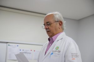 Médico Newton Tristão. Trata-se de um homem branco, de cabelos e bigode brancos. Ele usa óculos, jaleco branco e segura uma folha de papel com as mãos. Ao fundo, paredes na cor branca com papéis pregados em um quadro branco