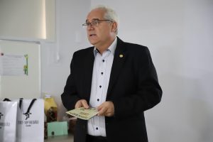 Desembargador Welington Peixoto. Trata-se de um homem branco de cabelos grisalhos. Ele usa óculos, paletó preto e camisa social branca. Ele está segurando a cartilha de prevenção ao assédio moral