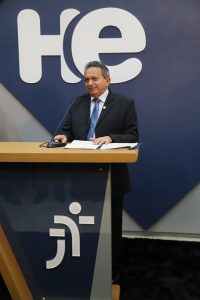 Foto colorida do desembargador Geraldo Nascimento, presidente do TRT-18. Trata-se de um homem de pele clara e cabelos grisalhos, posicionado atrás de um púlpito e falando ao microfone. Ele usa um paletó preto, uma camisa social branca e uma gravata azul.