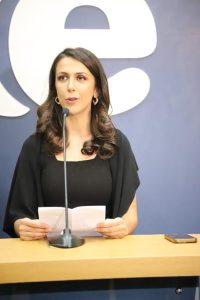 Foto colorida da coordenadora de Comunicação Social do TRT-18, Lídia Nercessian. Trata-se de uma mulher de pele clara e cabelos castanhos. Ela usa uma blusa preta e está atrás de um púlpito falando ao microfone.