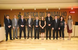 Sete homens, todos de terno, e três mulheres posam para foto lado a lado, em pé. Eles estão posicionados sob seis spots de lâmpadas amarelas. Ao fundo, se lê a expressão Justiça do Trabalho plotada em um painel de MDF
