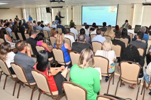 Uma plateia cheia, com dezenas de pessoas sentadas, assistem à exposição de um homem, que aponta para a tela em que estão projetados slides