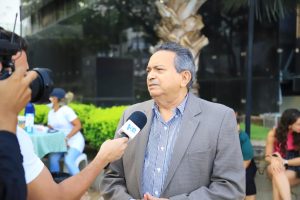 A imagem mostra um homem sendo entrevistado ao ar livre. Ele está sendo filmado por uma câmera enquanto segura um microfone de um repórter que está entrevistando-o. O homem está vestindo um blazer cinza sobre uma camisa listrada e parece estar respondendo a uma pergunta ou explicando algo. No plano de fundo, há algumas pessoas sentadas ao redor de mesas, indicando que a entrevista está ocorrendo em um local público como uma praça. A cena transmite uma atmosfera casual e diurna.